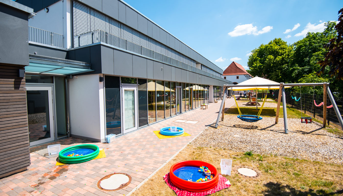 Spielbereich mit Planschbecken, Schaukeln und moderner Kita bei Sonnenschein