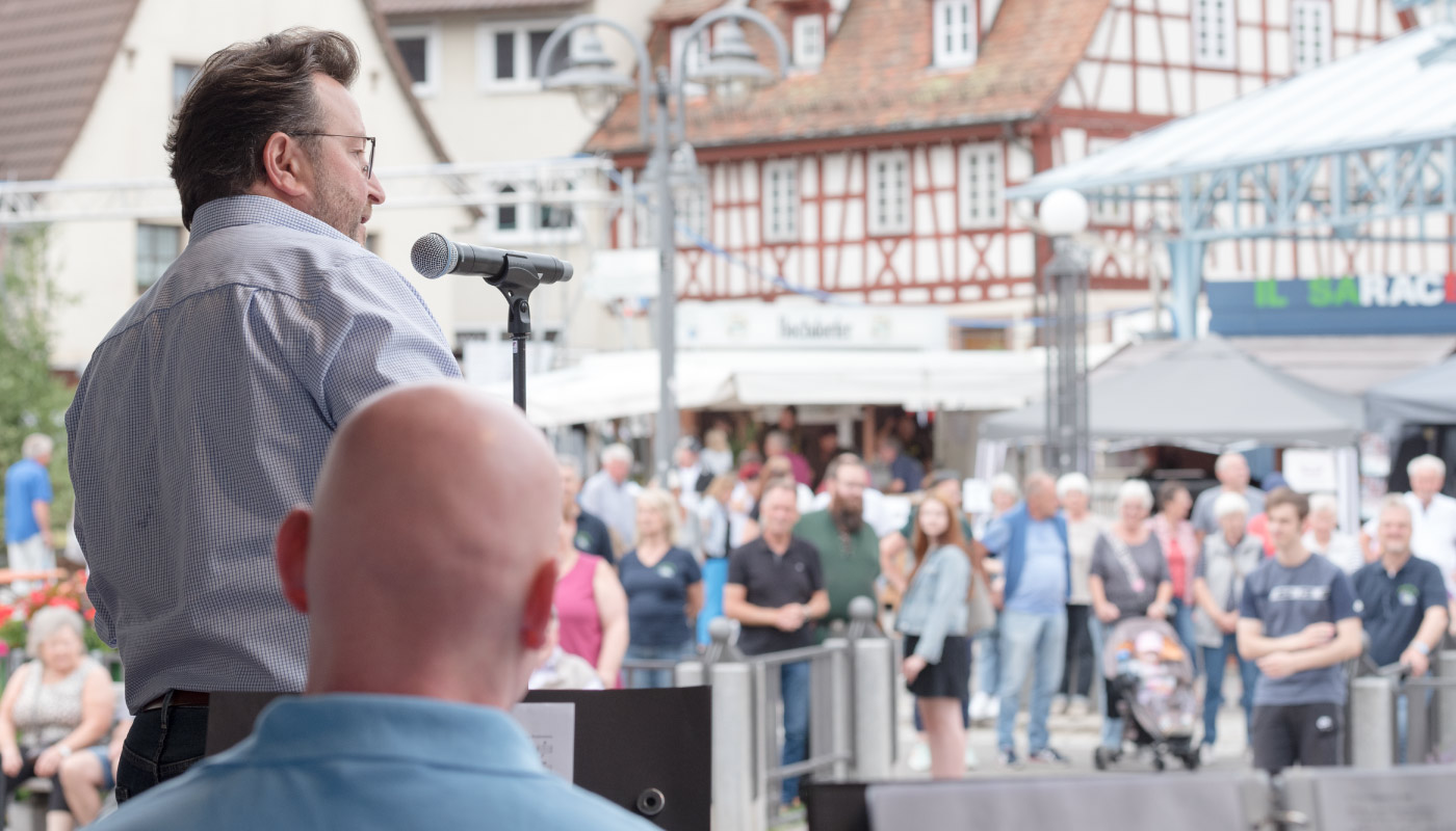 Mann spricht mit Mikrofon vor Publikum auf Marktplatz mit Fachwerkhäusern in Birkenfeld
