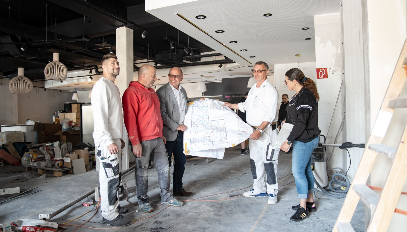 Fünf Personen stehen in einem renovierungsbedürftigen Innenraum und betrachten gemeinsam Baupläne